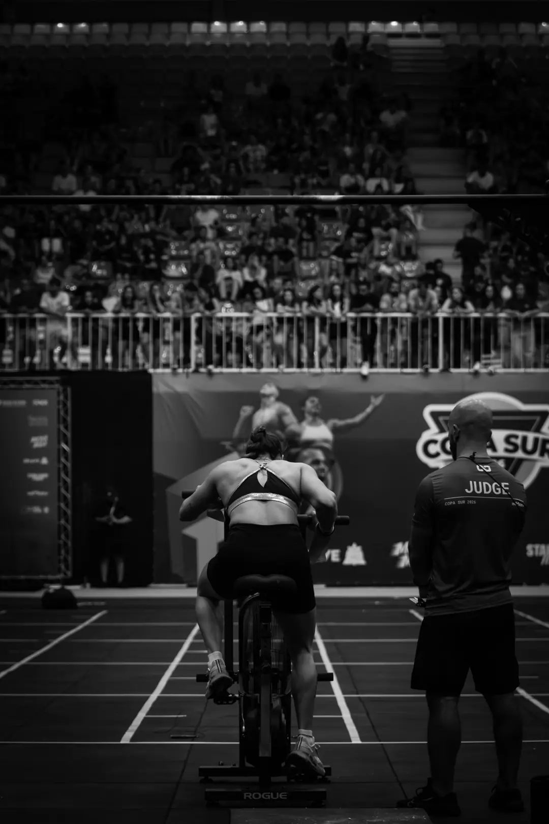 An athlete on an air bike at a competition, a judge standing beside her